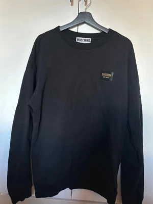 Svart sweatshirt från Moschino - Svart sweatshirt från Moschino med rund halsringning och diskret logopatch i svart och guld på bröstet. Tröjan har en klassisk passform och är tillverkad i mjukt bomullsmaterial. Perfekt för en stilren och trendig look.