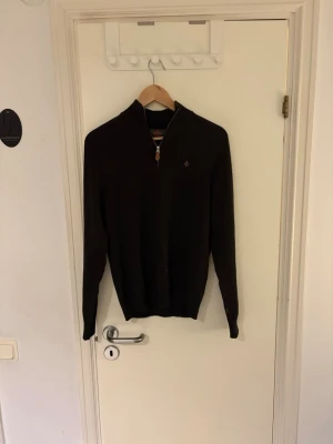 Brun halvzip-tröja från Morris - Snygg brun stickad tröja med halv dragkedja och liten broderad logga på bröstet. Tröjan har hög krage och långa ärmar, perfekt för lager-på-lager. Ribbstickade muddar vid ärmslut och nederkant ger en schysst passform.