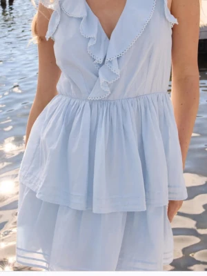 Ljusblå volangklänning med v-ringning - Söt ljusblå klänning med v-ringning och volangdetaljer framtill. Klänningen är ärmlös och har en luftig, kort modell med dubbla lager i kjolen. Perfekt för varma dagar och somriga tillfällen.