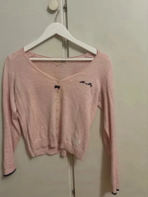 Ljusrosa cardigan Odd Molly - Söt ljusrosa cardigan från Odd Molly med v-ringning och knappar framtill. Liten rosett vid översta knappen och broderad logga på bröstet. Långärmad och kortare modell med ribbade muddar och diskret patch nertill.