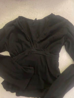 Svart omlottopp med knytband - Svart omlottopp med v-ringning och långa ärmar. Toppen har ett brett knytband i midjan. Perfekt att styla med jeans eller kjol för en trendig look.