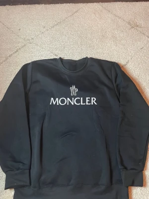 Moncler sweatshirt  - Snygg svart sweatshirt från Moncler med vit logotyp och text på bröstet. Tröjan har rund halsringning, långa ärmar och ribbade muddar vid ärmslut och nederkant. Perfekt för en clean och stilren look.