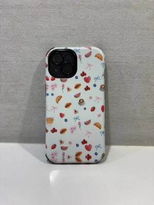 Vitt mobilskal med frukt- och fikamotiv - Snyggt mobilskal i vitt med färgglada motiv av frukter, bär, croissanter och små rosetter. Skalet har en matt finish och är gjort i tålig plast med svart insida. Perfekt för dig som vill ha ett lekfullt och unikt skydd till din mobil. Helt nytt, oanvänt. Till iPhone 15.