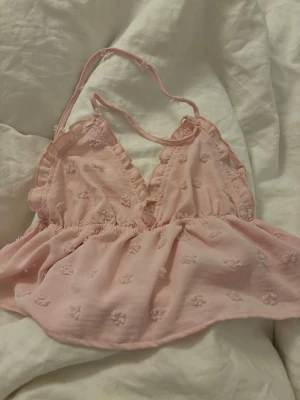 Ljusrosa babydoll med broderade blommor - Söt ljusrosa babydoll med tunna axelband och volangkant vid kuporna. Tyget är dekorerat med små broderade blommor och har en lätt, luftig känsla. Perfekt för dig som gillar romantiska och feminina detaljer.