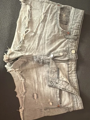 Grå slitna jeansshorts med knappar - Säljer ett par grå jeansshorts med slitna detaljer och råa kanter. Shortsen har knappgylf, fem fickor och en cool, avslappnad look. Perfekta för dig som gillar en edgy stil.