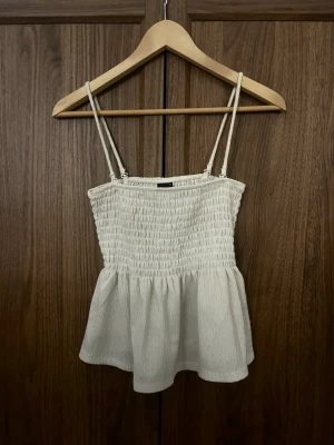 Vit linnetopp Gina Tricot - Supersöt vit linnetopp från Gina Tricot med smala axelband och smockad överdel. Toppen har en luftig, lätt utställd nederdel och är perfekt för varma dagar. Materialet känns mjukt och bekvämt mot huden.