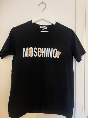 Svart Moschino t-shirt med björnar - Snygg svart t-shirt från Moschino med stort vitt tryck på bröstet där det står MOSCHINO och två gulliga björnar som detaljer. Klassisk rund halsringning och korta ärmar. Perfekt statement-plagg för dig som gillar streetstyle och designer.