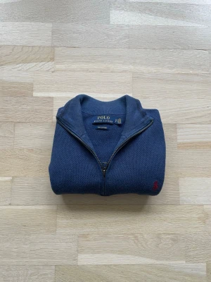 Blå halvzip-tröja från Polo Ralph Lauren - Mörkblå stickad halvzip-tröja från Polo Ralph Lauren i bomull. Tröjan har en klassisk röd broderad logga på bröstet och grovstickad struktur. Perfekt för lager-på-lager och snygg till jeans eller chinos.