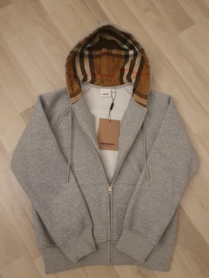 Grå zip hoodie från Burberry - Snygg grå hoodie från Burberry med dragkedja och klassisk beige, brun och svart rutig huva. Hoodien har ribbade muddar och dragsko i huvan. Tillverkad i mjuk bomull för maximal komfort. Perfekt för en avslappnad och trendig look.