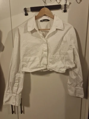 Vit croppad skjorta från Zara - Säljer en vit croppad skjorta från Zara  med klassisk krage och långa ärmar. Skjortan har knappar framtill och två fickor på bröstet. Perfekt för en trendig och avslappnad look.