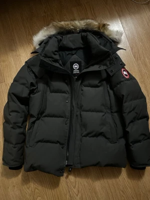 Grå dunjacka från Canada Goose - Säljer en grå dunjacka från Canada Goose med avtagbar huva och snygg pälskant. Jackan har klassisk patch på ärmen, dragkedja och knappar framtill samt stora fickor. Perfekt för kalla vinterdagar och riktigt varm. 1:1