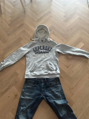Grå hoodie från Superdry med tryck - Snygg grå hoodie från Superdry med blått och vitt tryck på bröstet och ärmen. Hoodien har en mysig fodrad huva och  känguruficka. Perfekt för en avslappnad och sportig stil. Materialet är mjukt och bekvämt, perfekt för vardagslooken. Skriv vid minds fundering och pris kan diskuteras!
