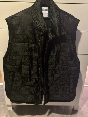 Svart dunväst från Moschino - Snygg svart dunväst från Moschino med logotypmönster över hela västen. Västen har hög krage, dragkedja och stora fickor framtill. Perfekt för att lyfta din streetwear-look med en exklusiv touch.