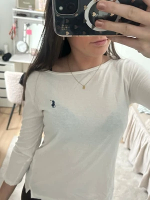 Vit långärmad topp från Ralph Lauren - Snygg vit trekvart tröja från Ralph Lauren med klassisk marinblå logga broderad på bröstet. Lite större vid halsen, men så modellen ska vara. perfekt att styla med jeans eller kjol. Mjukt och bekvämt material.