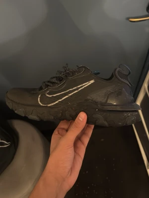 Svarta Nike React Element sneakers - Säljer ett par svarta Nike React Element sneakers med vit swoosh på sidan. Skorna har en chunky sula med vågig design och reflekterande detaljer på snörena. Ovandelen är i mesh och syntetmaterial för bra andningsförmåga och komfort.