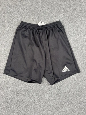 Svarta träningsshorts från Adidas - Säljer ett par svarta träningsshorts från Adidas med klassisk logga på vänster ben. Shortsen har elastisk midja och är tillverkade i ett lätt och snabbtorkande syntetmaterial, perfekta för sport och aktivitet.