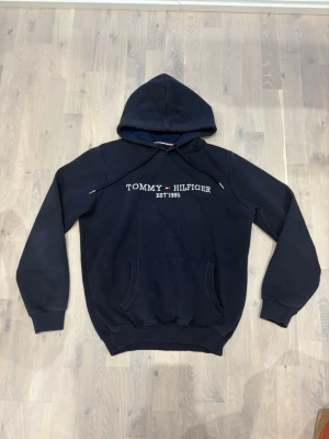 Svart hoodie från Tommy Hilfiger - Snygg svart hoodie från Tommy Hilfiger med broderad logga och text 'EST. 1985' på bröstet. Klassisk känguruficka framtill, justerbar huva med dragsko och ribbade muddar vid ärmar och nederkant. Perfekt för en avslappnad och stilren look.