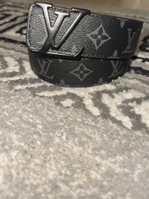  Svart monogram bälte i skin - Snyggt svart skärp med klassiskt monogrammönster och stor logga i metall som spänne. Skärpet är gjort i skinn och har en stilren design som passar till många outfits.