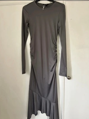 Grå långärmad långklänning - Snygg grå långklänning med lång ärm och asymmetrisk volangkant nedtill. Klänningen har en lätt rynkning i sidan som ger en cool detalj och är tillverkad i ett mjukt, stretchigt material. Perfekt för dig som gillar minimalistisk och stilren design.