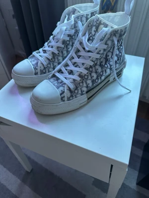 Dior B23 high-top sneakers - Säljer ett par Dior B23 high-top sneakers med det ikoniska monogrammönstret i grått och vitt. Skorna har vita snören, genomskinliga paneler och robust vit gummisula. Perfekt för dig som vill sticka ut med lyxig streetwear.