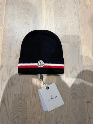 Svart Moncler mössa - Snygg svart ribbstickad mössa från Moncler med uppvikt kant. Mössan har en broderad Moncler-logga framtill och en randig detalj i rött, vitt och blått längs kanten. Perfekt accessoar för att lyfta din stil under kyliga dagar.