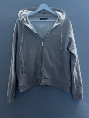 Beige hoodie (Lexington) - Snygg grå hoodie från Lexington med dragkedja framtill och huva med dragsko. Diskret Lexington-logga på bröstet och två fickor på sidorna. Perfekt för en chill och avslappnad stil.