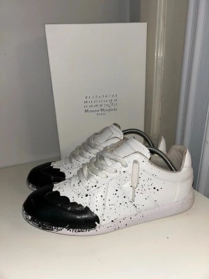 Maison Margiela svartvita sneakers - Unika sneakers från Maison Margiela i vitt läder med svarta detaljer och färgstänk. Låg modell med klassisk snörning och gummisula. Snygg kontrast mellan svart och vitt som ger en cool och modern vibe.