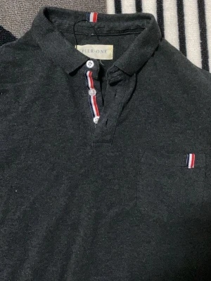 Svart henley från Pier One - Svart henley från Pier One med krage och knappar framtill. Snygga detaljer med röd, vit och blå rand längs knappslån och på bröstfickan. Skön bomullskvalitet och klassisk passform, perfekt till jeans eller chinos.