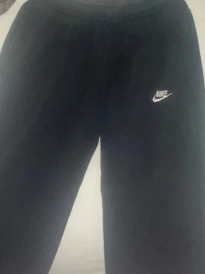 Svarta mjukisbyxor från Nike - Svarta mjukisbyxor från Nike med klassisk vit logga på vänster ben. Byxorna har en avslappnad passform och resår i midjan. Perfekta för chill eller träning. Mjukt material som känns skönt mot huden.