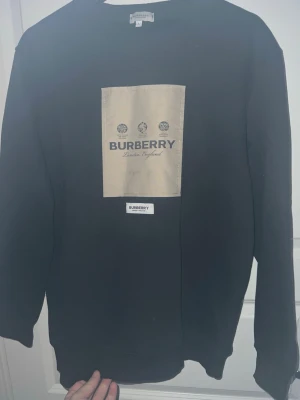 Svart sweatshirt från Burberry - Stilren svart sweatshirt från Burberry med beige patch och logga på bröstet. Klassisk rund halsringning och långa ärmar. Perfekt för dig som gillar exklusiva streetwear-plagg med tydlig branding.