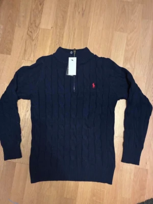 Mörkblå kabelstickad tröja Polo Ralph Lauren - Helt ny Mörkblå kabelstickad tröja från Polo Ralph Lauren med halv dragkedja och röd broderad logga på bröstet. Tröjan har ribbade muddar och klassisk passform, perfekt för lager-på-lager. Materialet ser ut att vara mjukt och värmande. Lapparna är fortfarande på, ett till ett , fraktar snabbt, storleken är M men skulle säga att den passar också bra på S