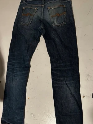 Mörkblå jeans från Nudie Jeans - Säljer två par jeans från Nudie Jeans med klassisk femficksdesign och snygga kontrastsömmar på bakfickorna. Jeansen har en rak passform och är tillverkade i denim med lätt slitning för en cool look. De har blivit andvända ett tag men ser jävligt bra ut. De mörk  blå är modell steady Eddie medans de andra är Grim Tim. Har några slitningar men gör det hela bättre.