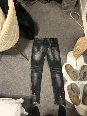 Svarta skinny jeans med slitningar - Säljer ett par svarta skinny jeans med kraftiga slitningar och vita färgstänk. Jeansen har en tvättad look, detaljerade slitningar framtill och klassiska bakfickor. Perfekta för dig som gillar en edgy och trendig stil.