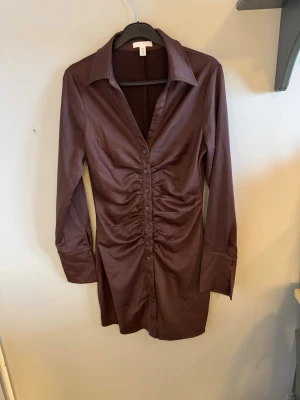 Brun skjortklänning med knappar - Snygg brun skjortklänning med långa ärmar och knappar framtill. Klänningen har en figurnära passform med rynkade detaljer längs knappraden och klassisk krage. Perfekt för dig som gillar stilrena och trendiga plagg.
