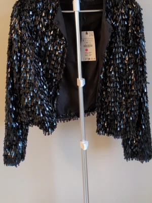 Svart paljettkavaj från Zara - Glittrig svart kavaj från Zara täckt med glänsande paljetter. Kort modell med lång ärm och öppen front med små hakar till knäppning. Perfekt för dig som vill sticka ut och addera extra bling till din outfit. Fodrad insida för skön känsla. Prislappen kvar.