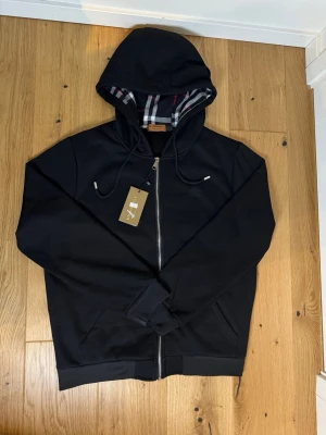 Burberry Hoodie - Snygg svart hoodie från Burberry med klassiskt rutigt foder i huvan och dragkedja framtill. Broderad logga på bröstet och justerbar huva med snören. Perfekt för en stilren och trendig look.