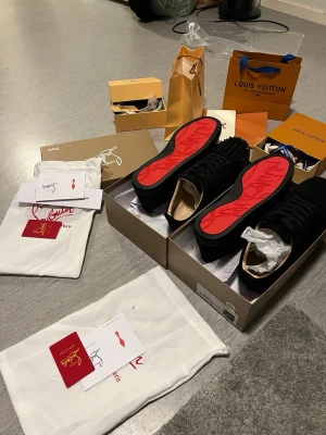 Svarta Louboutin sneakers med röd sula - Säljer ett par svarta sneakers från Christian Louboutin med ikonisk röd sula och svarta snören. Ovandelen är i mocka och har coola nitar på tån för extra edge. Skorna kommer med originalkartong och dustbag. Perfekta för dig som vill sticka ut.