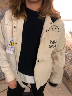 Beige overshirt med smiley-tryck - Beige overshirt med svart tryck på bröstfickan där det står 'Smile Today' och en handritad smiley. Jackan har knappar framtill och en avslappnad passform. Perfekt för lager-på-lager och streetstyle.