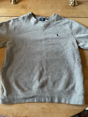 Grå sweatshirt från Polo Ralph Lauren - Klassisk grå sweatshirt från Polo Ralph Lauren med rund halsringning och diskret broderad logga på bröstet. Tröjan har ribbade muddar vid ärmslut och nederkant. Perfekt för en avslappnad och stilren look.