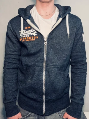 Mörkgrå zip-hoodie från Superdry - Mörkgrå hoodie med dragkedja från Superdry. Tryck i vitt och orange på bröstet samt vitt tryck på ärmen. Huva med vita snören och två fickor framtill. Mjuk bomullsblandning och sportig look.