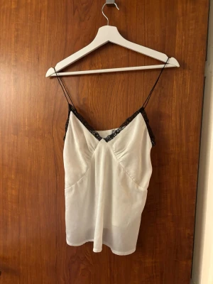 Vit linnetopp med spetsdetaljer - Säljer en vit linnetopp med tunna axelband och svart spets längs urringningen. Toppen har en mjuk, glansig yta som påminner om satin och en feminin v-ringad form framtill. Perfekt att styla lager på lager eller bära som den är.