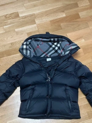 Svart jacka  från Burberry - Säljer en svart jacka  från Burberry med avtagbara ärmar och klassiskt rutigt foder i huvan. Västen har två dragkedjeförsedda fickor framtill och en broderad Burberry-logga på ena ärmen. Perfekt för dig som vill ha en stilren och lyxig look.