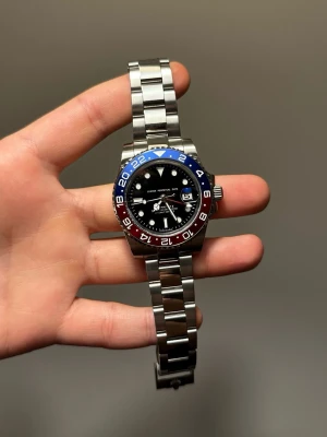 Stålarmbandsklocka med blå/röd bezel - Snygg klocka med svart urtavla, datumvisning och tvåfärgad blå/röd bezel. Armbandet är i rostfritt stål och klockan har tydliga indexmarkeringar samt vridbar ring. Perfekt accessoar för dig som gillar sportig stil.