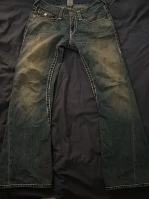 True Religion bootcut jeans blå - Säljer ett par blå bootcut jeans från True Religion med slitna detaljer och kontrastsömmar. Jeansen har klassiska fickor med lock bak och tydliga broderier. Perfekta för dig som gillar en avslappnad och trendig stil.
