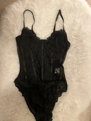 Svart spetsbody med tunna axelband - Snygg svart body i spets med tunna justerbara axelband och dekorativa banddetaljer framtill. Bodyn har vacker spetskant vid bysten och transparenta partier längs sidorna. Perfekt för dig som vill ha en elegant och trendig look.