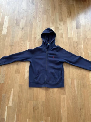 Mörkblå hoodie från Polo Ralph Lauren - Snygg mörkblå hoodie från Polo Ralph Lauren med dragkedja framtill och klassisk huva. Tröjan har två fickor på magen och den ikoniska röda loggan broderad på bröstet. Tillverkad i mjukt bomullsmaterial som känns skönt mot huden.