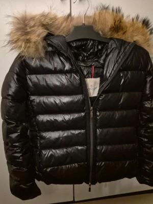 Svart dunjacka med pälskrage Moncler - Snygg svart dunjacka från Moncler med glansig finish och stor huva med fluffig pälskant. Jackan har quiltad design, dragkedja framtill och en klassisk pufferstil. Jackan har några år på nacken men fint skick för övrigt.