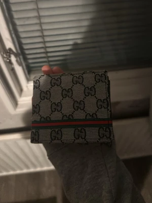 Gucci GG Supreme plånbok grå - Snygg grå plånbok från Gucci med klassiskt GG Supreme-mönster och en röd-grön rand på framsidan. Plånboken har flera fack för kort och sedlar samt ett genomskinligt fönster för ID. Tillverkad i canvas och läder.