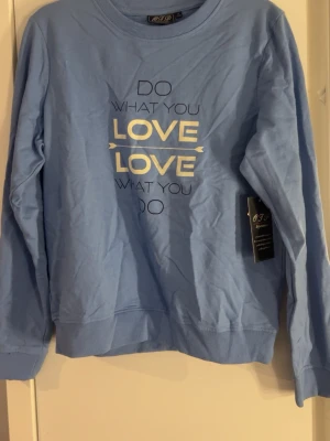 Ljusblå sweatshirt med tryck - Ljusblå sweatshirt från GFJ med texten 'DO WHAT YOU LOVE LOVE WHAT YOU DO' i vitt och svart på bröstet. Tröjan har rund hals, långa ärmar och ribbade muddar vid ärmslut och nederkant. Perfekt för en avslappnad och trendig look.