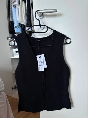 Svart  väst med knappar - Stilren svart väst i modell med v-ringning och knappar framtill. Perfekt att bära över en skjorta eller t-shirt för en trendig lager-på-lager-look. Västen har en klassisk passform och är enkel att matcha med olika outfits. Passar som xs/s. 